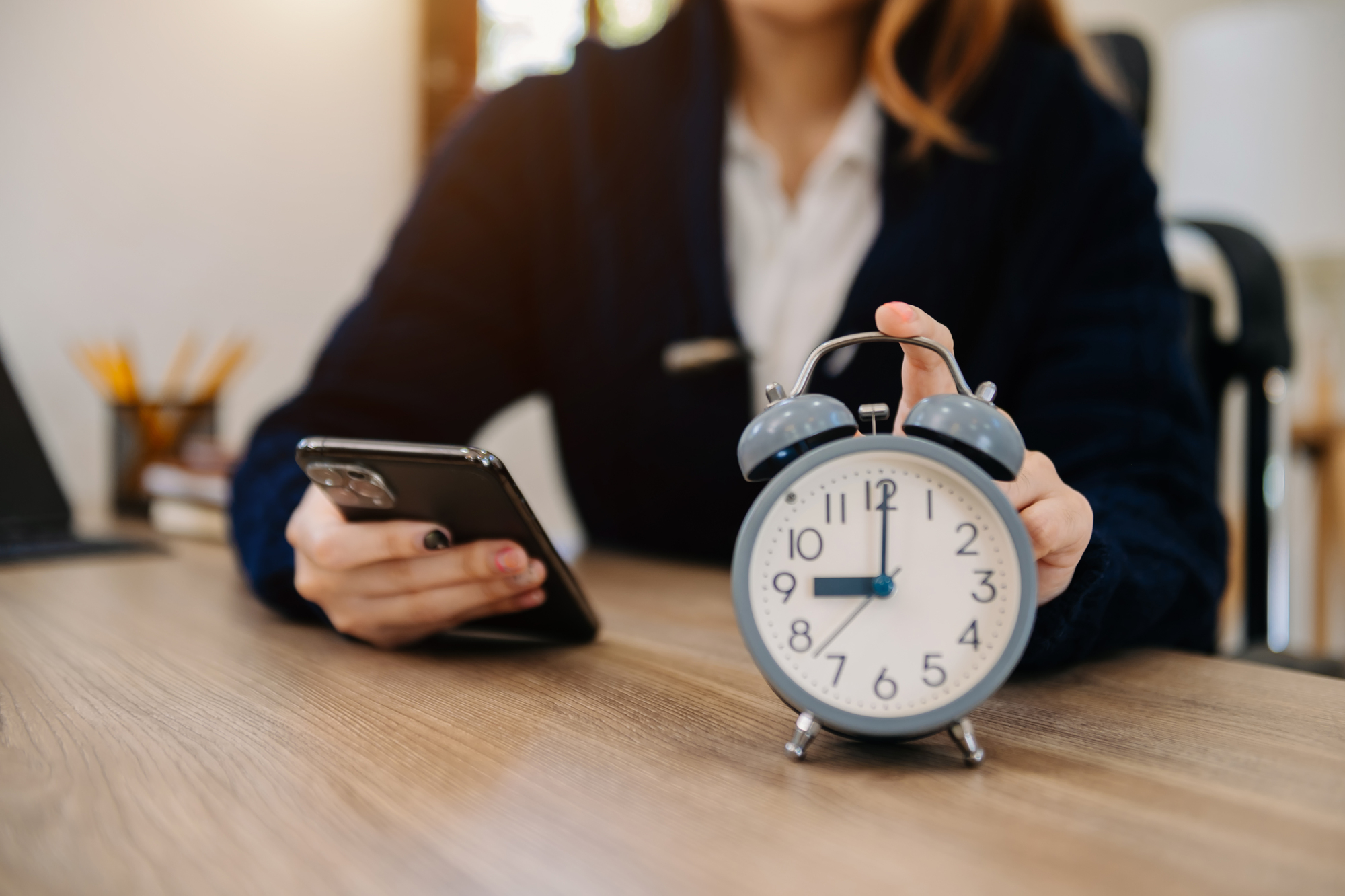 A person sits at a desk holding a smartphone in one hand and touching the top of a classic alarm clock with the other hand. The alarm clock is placed on the desk, showing the time as 7:00. The background includes blurred office supplies.