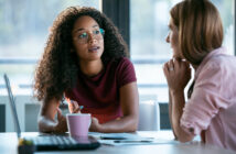 Two women are engaged in a conversation at an office table. One woman with curly hair and glasses is speaking, holding a pen. The other woman, with straight hair, listens attentively with her chin resting on her hand. A laptop and pink mug are on the table.