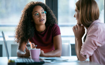 Two women are engaged in a conversation at an office table. One woman with curly hair and glasses is speaking, holding a pen. The other woman, with straight hair, listens attentively with her chin resting on her hand. A laptop and pink mug are on the table.