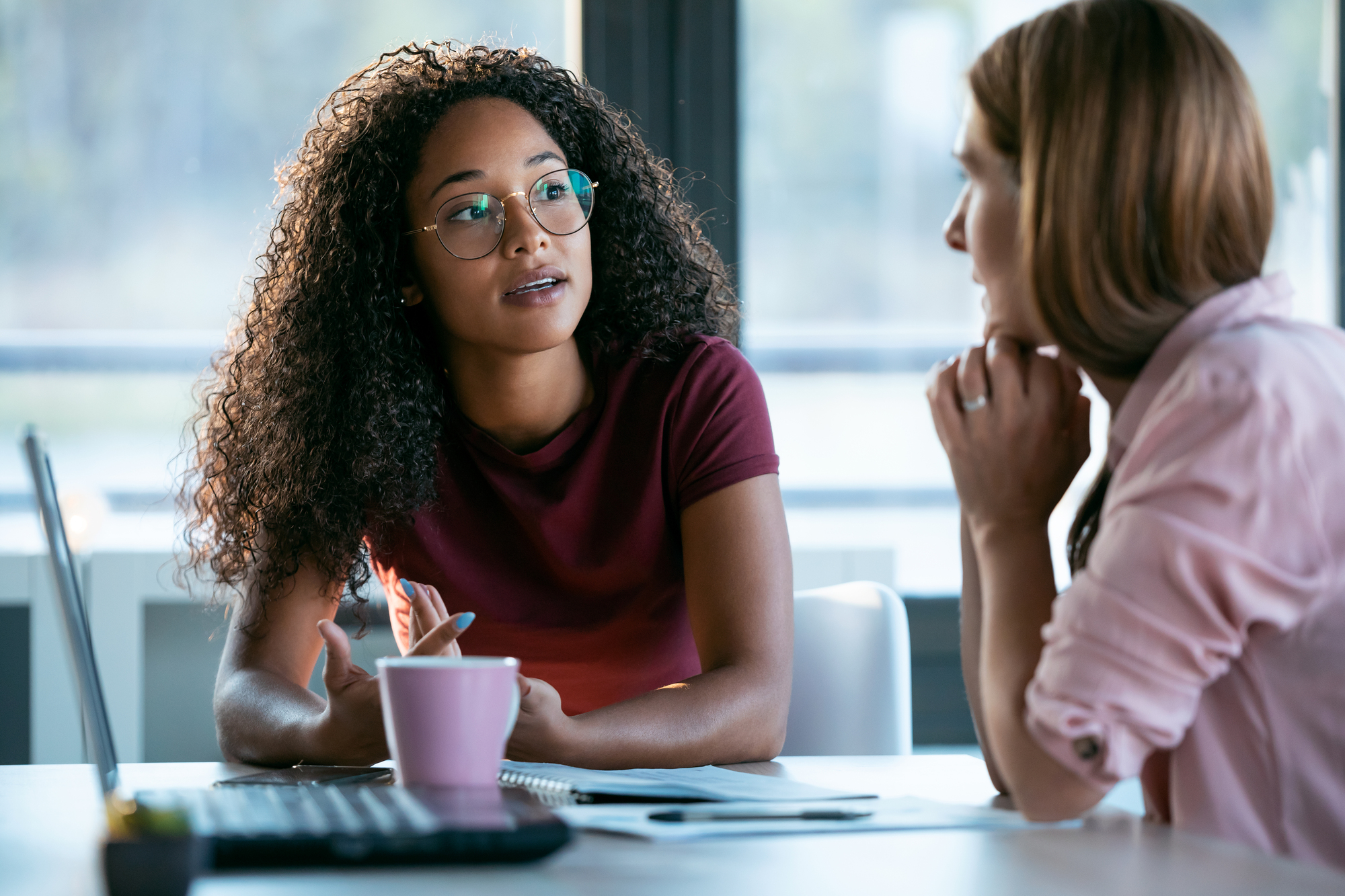 Two women are engaged in a conversation at an office table. One woman with curly hair and glasses is speaking, holding a pen. The other woman, with straight hair, listens attentively with her chin resting on her hand. A laptop and pink mug are on the table.