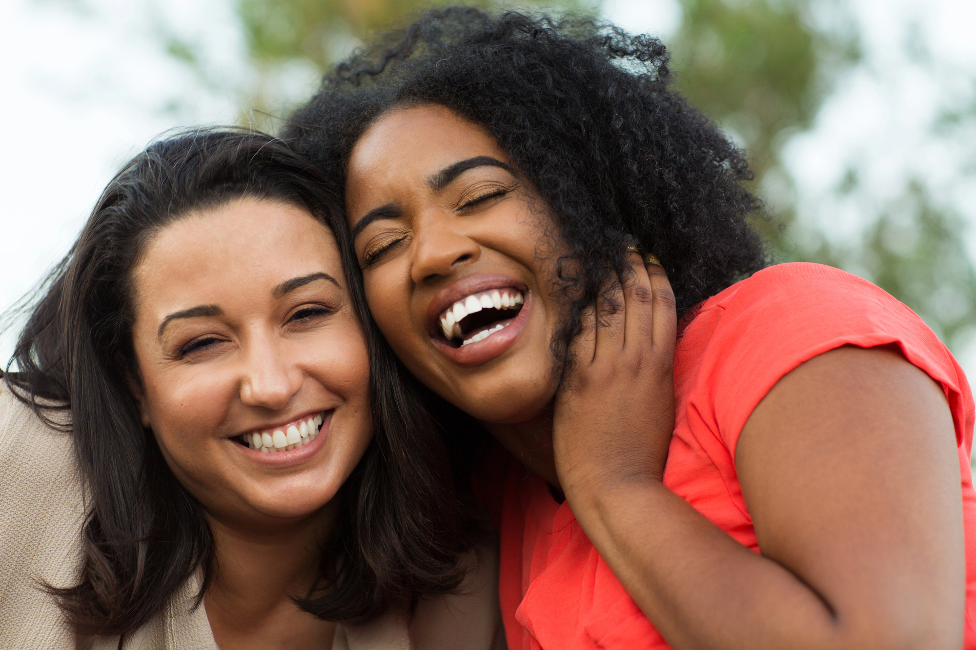 Two women are smiling and laughing outdoors. One woman has shoulder-length dark hair, and the other has curly hair, wearing a red top. They appear happy and are leaning close to each other, with a blurred background of greenery.