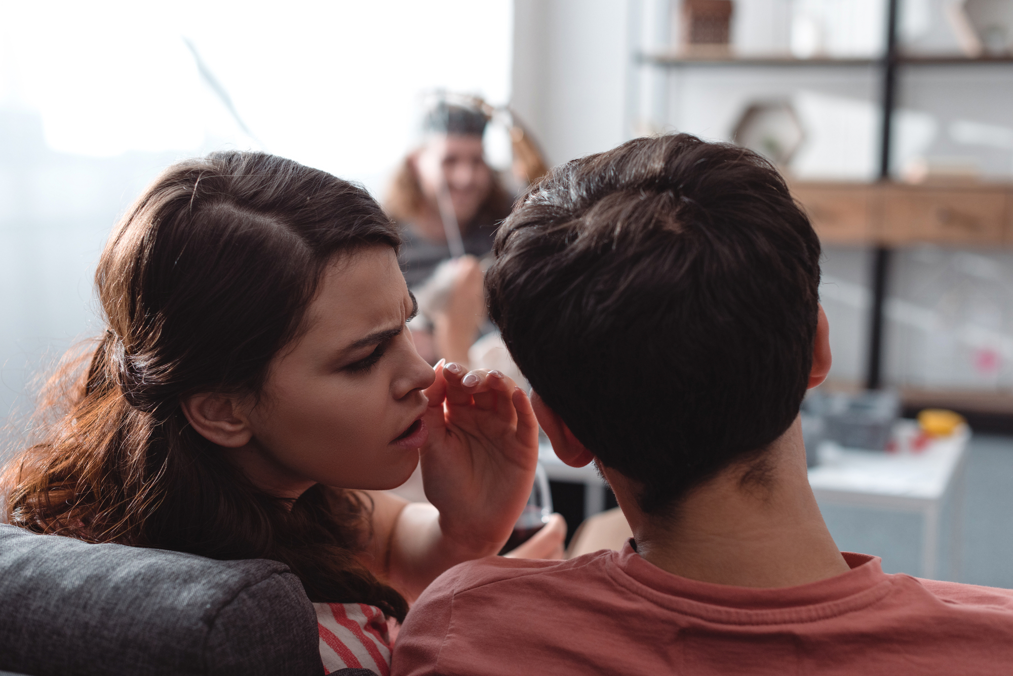 Two people are sitting on a couch, with one whispering into the other's ear. In the background, a person is sitting at a desk, wearing headphones and smiling. Shelves and household items are visible in the softly lit room.