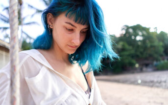 A person with blue hair and multiple earrings is standing outdoors, looking downwards. They are wearing a white shirt over a black top. The background features a beach with trees and structures. The lighting suggests it's either morning or late afternoon.