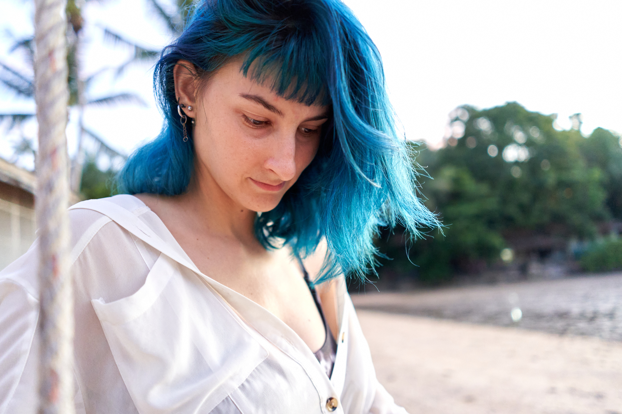 A person with blue hair and multiple earrings is standing outdoors, looking downwards. They are wearing a white shirt over a black top. The background features a beach with trees and structures. The lighting suggests it's either morning or late afternoon.