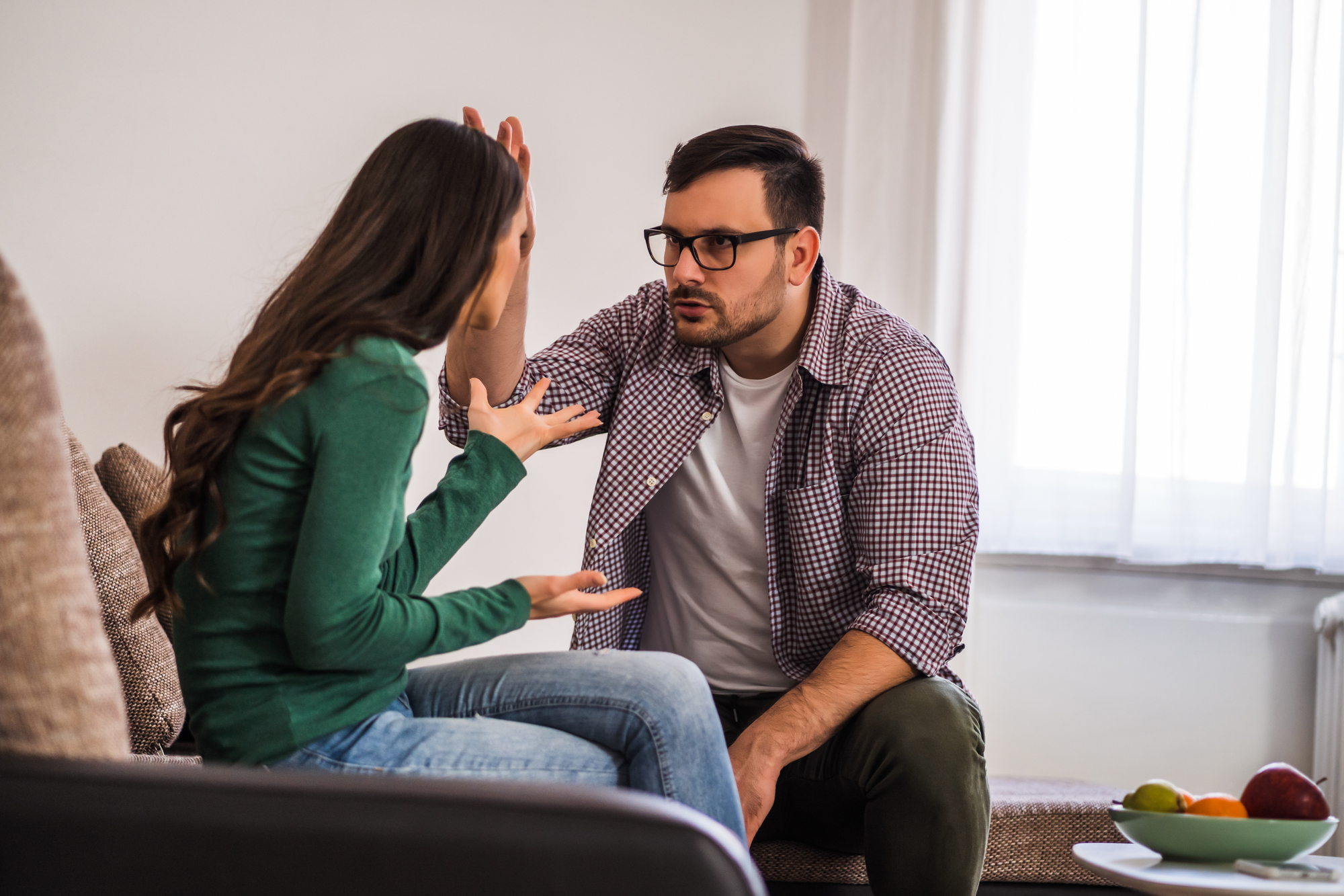 A woman and a man are having an intense conversation while sitting on a couch in a living room. The woman is gesturing with her hands, and the man, wearing glasses and a checkered shirt, is leaning forward and looking at her seriously. A fruit bowl is visible on a nearby table.