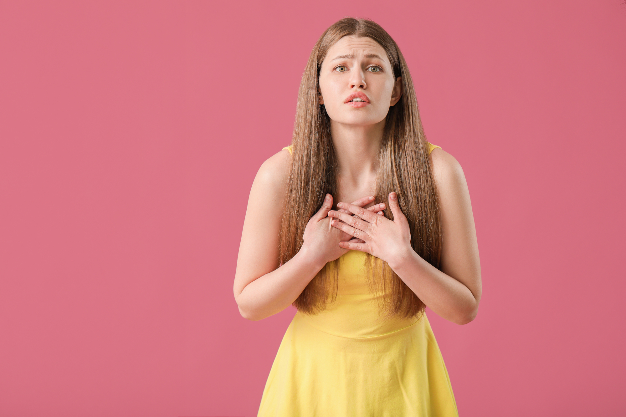 A woman with long brown hair stands against a pink background. She is wearing a sleeveless yellow dress and has a concerned expression on her face, with both hands placed on her chest.