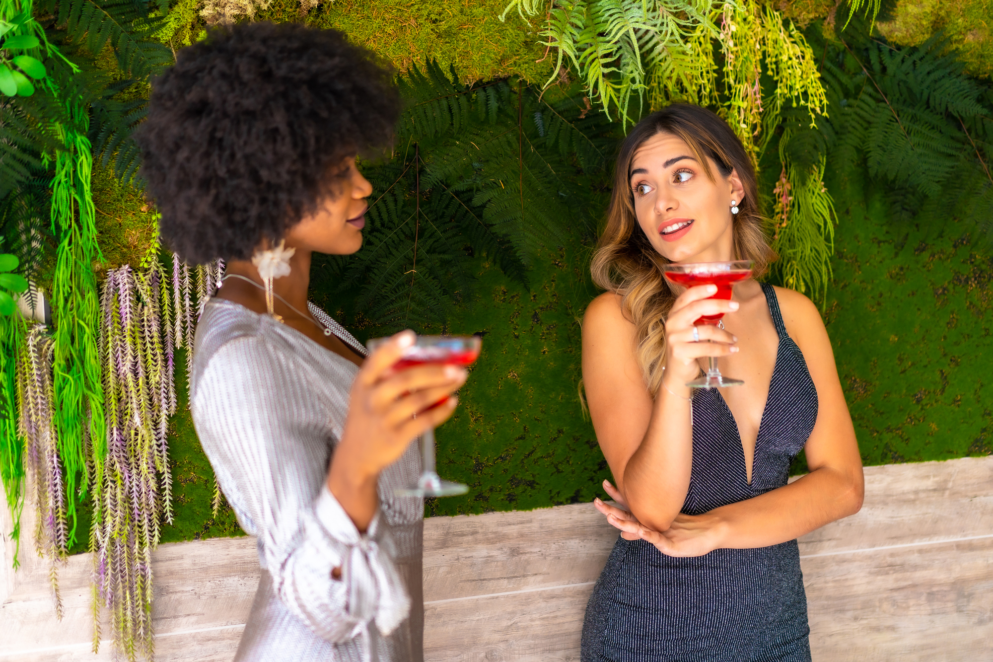 Two women stand smiling and holding red cocktails against a backdrop of lush greenery and foliage. One has curly, short hair and wears a shimmering long-sleeve dress, while the other has long, wavy hair and wears a sleeveless dark dress.