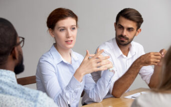 A woman with red hair is speaking and gesturing with her hands during a meeting. She is seated at a table with three colleagues, who are listening attentively. They are all wearing business casual attire and have papers in front of them.