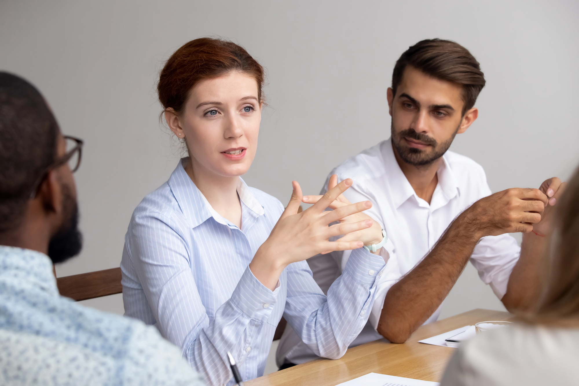 A woman with red hair is speaking and gesturing with her hands during a meeting. She is seated at a table with three colleagues, who are listening attentively. They are all wearing business casual attire and have papers in front of them.