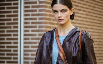 A person with brown hair neatly pulled back and parted in the middle is wearing a brown leather jacket over a blue knitted top. They are carrying a brown shoulder bag and standing in front of a brick wall background. They have a serious facial expression.