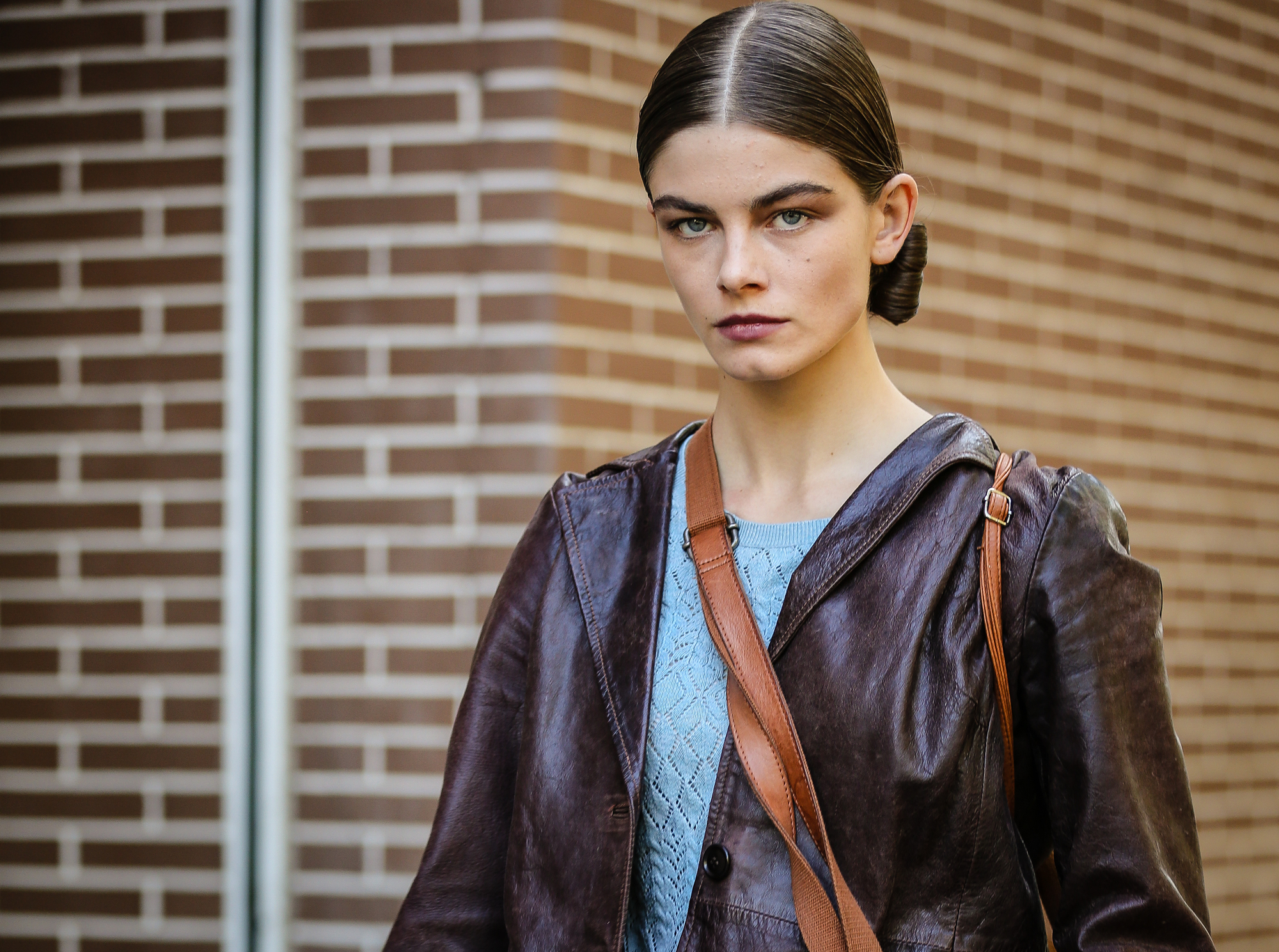 A person with brown hair neatly pulled back and parted in the middle is wearing a brown leather jacket over a blue knitted top. They are carrying a brown shoulder bag and standing in front of a brick wall background. They have a serious facial expression.