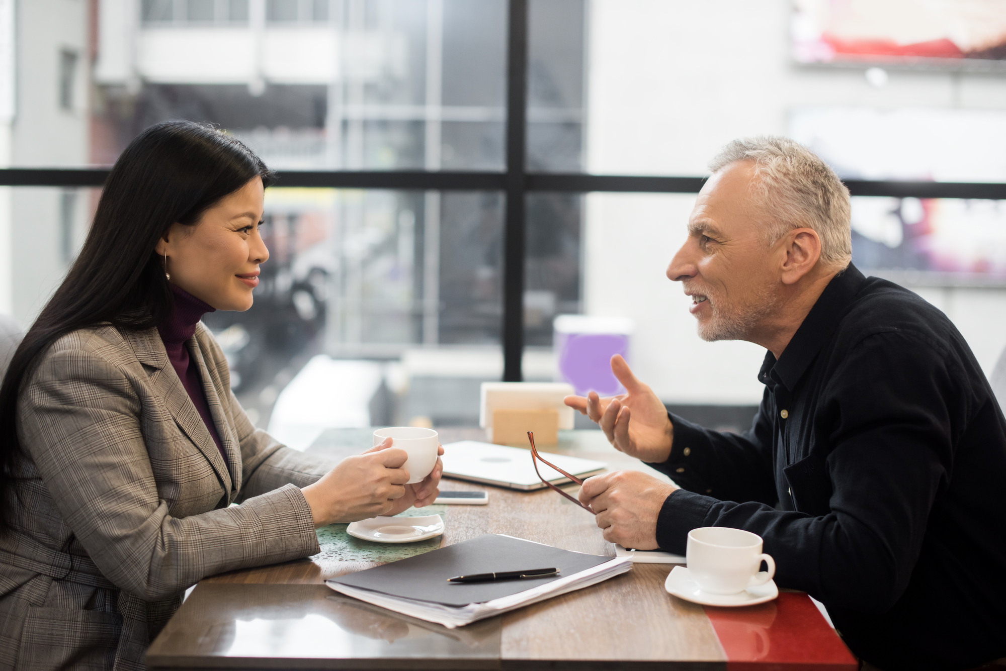 A woman and a man sit across from each other at a table in a modern café, engaged in conversation. Both have coffee cups in front of them, along with papers and a tablet. The woman holds her cup, and the man gestures with his hands while holding his glasses.