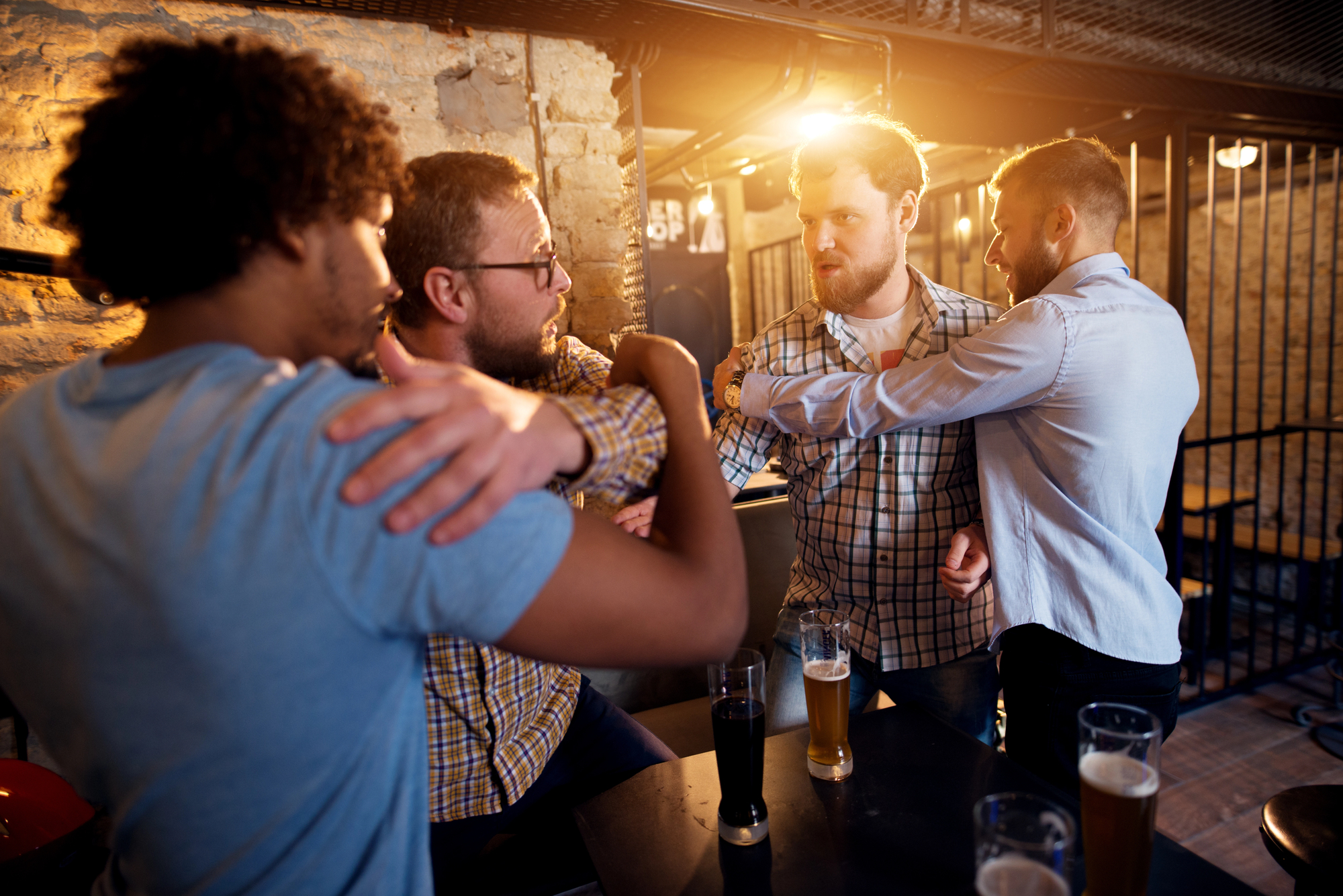 Four men in a bar are engaged in a heated argument. Two men, one in a plaid shirt and the other in a light blue shirt, are being held back by their friends. Glasses of beer are on the table in the foreground, and the scene is tense, with dramatic lighting.