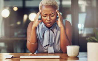 A person with short blonde hair sits at a desk with closed eyes, pressing their fingers to their temples in frustration. They are wearing a sleeveless top and have a coffee mug in front of them. The background is a dimly lit office space.