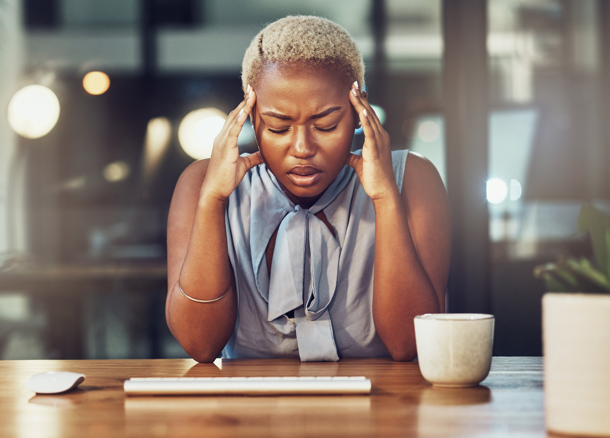 A person with short blonde hair sits at a desk with closed eyes, pressing their fingers to their temples in frustration. They are wearing a sleeveless top and have a coffee mug in front of them. The background is a dimly lit office space.