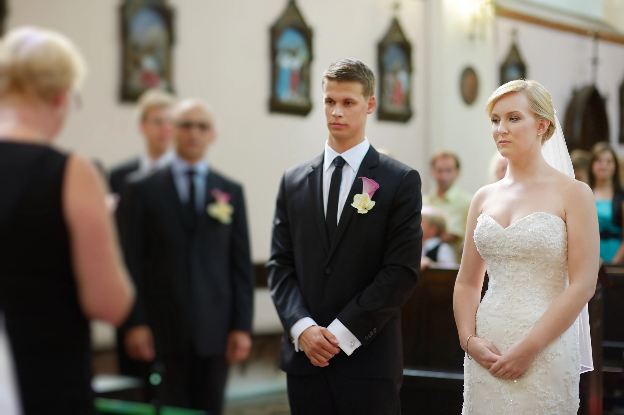 A bride and groom stand side by side in formal attire during a wedding ceremony in a church. The groom is in a black suit with a white shirt and tie, and the bride is in a white strapless wedding dress. A woman with short blonde hair, possibly officiating the ceremony, is in the foreground.