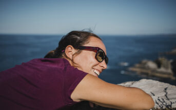 A woman wearing sunglasses and a purple shirt leans on a rock ledge, smiling while facing the ocean on a sunny day. She has her hair tied back and is enjoying the scenic view. The background features a clear blue sky and calm ocean waves.