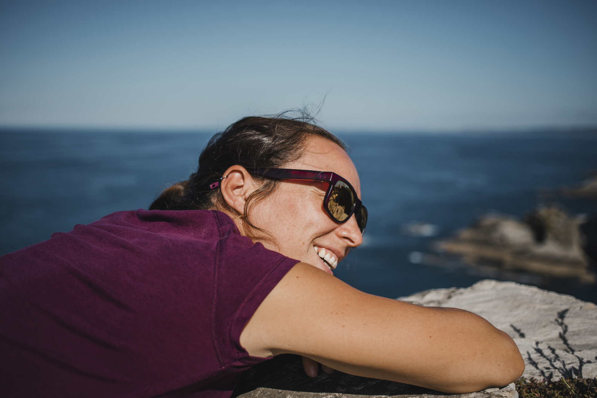 A woman wearing sunglasses and a purple shirt leans on a rock ledge, smiling while facing the ocean on a sunny day. She has her hair tied back and is enjoying the scenic view. The background features a clear blue sky and calm ocean waves.