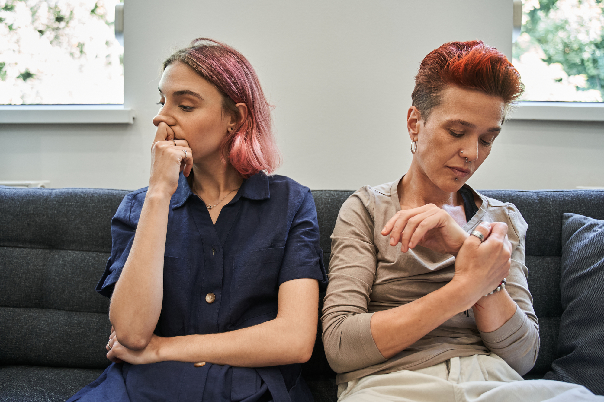 Two people are sitting on a couch, appearing to be in a serious or tense conversation. The person on the left, with pink hair, is looking to the side with a hand resting on their chin. The person on the right, with short red hair, is looking at their watch.