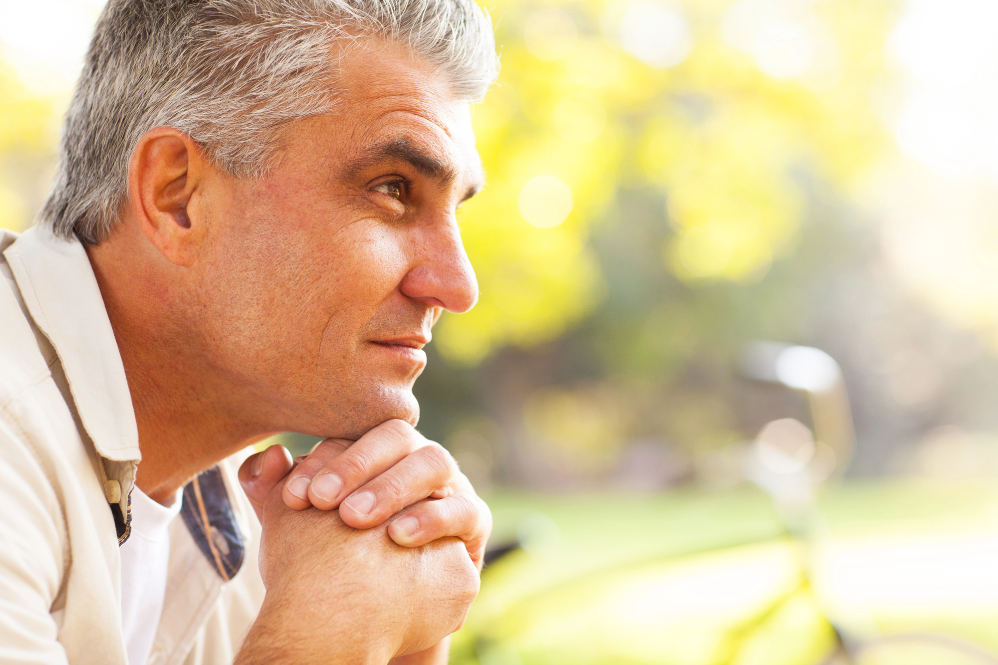 A man with gray hair is seated outdoors, resting his chin on his clasped hands. He looks to the right with a contemplative expression. The background is blurred, showing green foliage and a bicycle. The scene is brightly lit with natural sunlight.