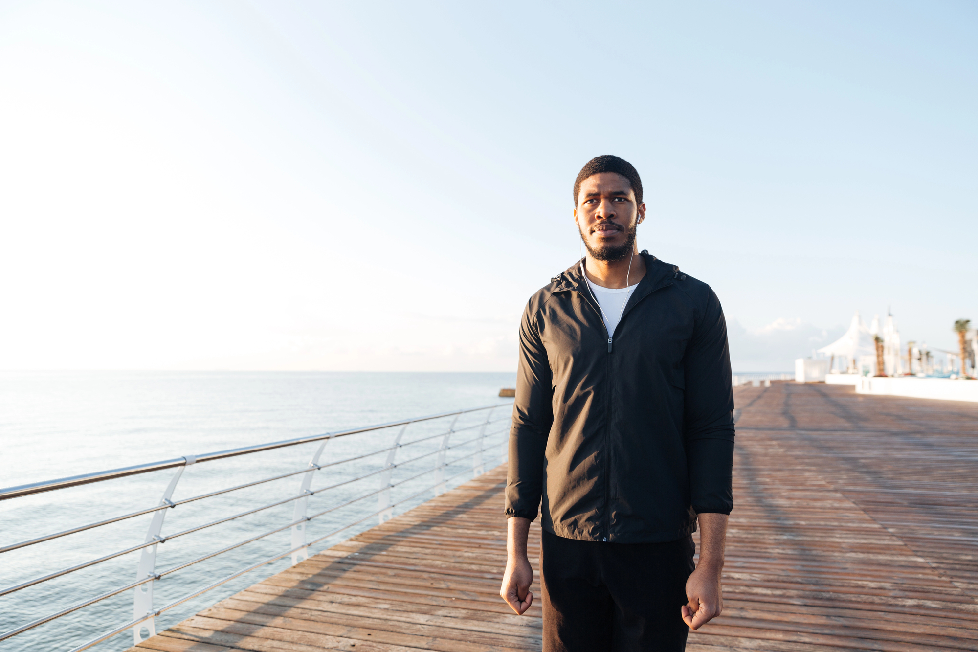 A man in a black jacket stands on a wooden boardwalk by the sea, with a railing to his left and a clear sky in the background. The boardwalk stretches into the distance.