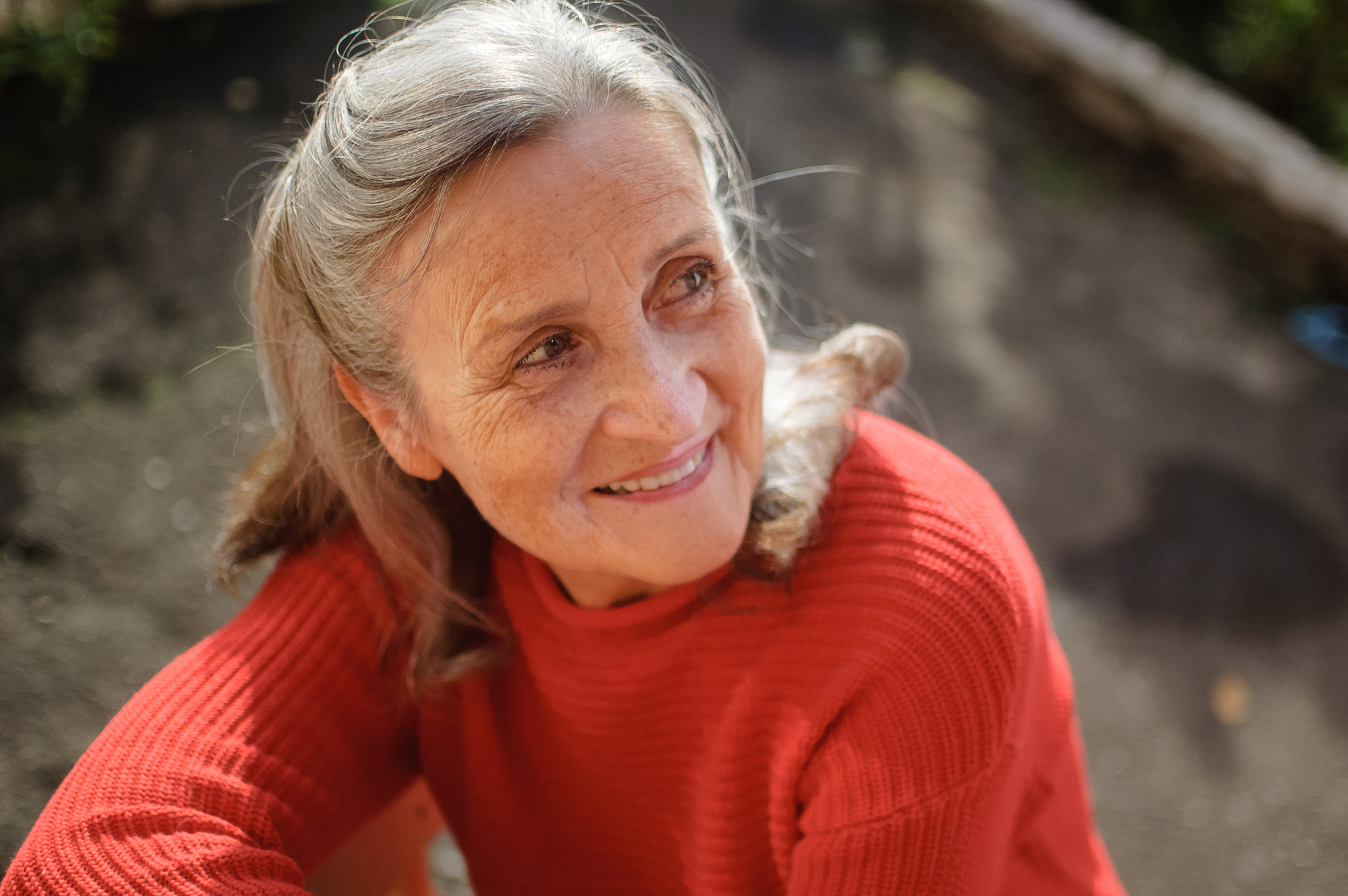 An elderly woman with gray hair, wearing a bright red sweater, smiles gently while looking away from the camera. She is outdoors with an unfocused background, possibly a garden or park setting.