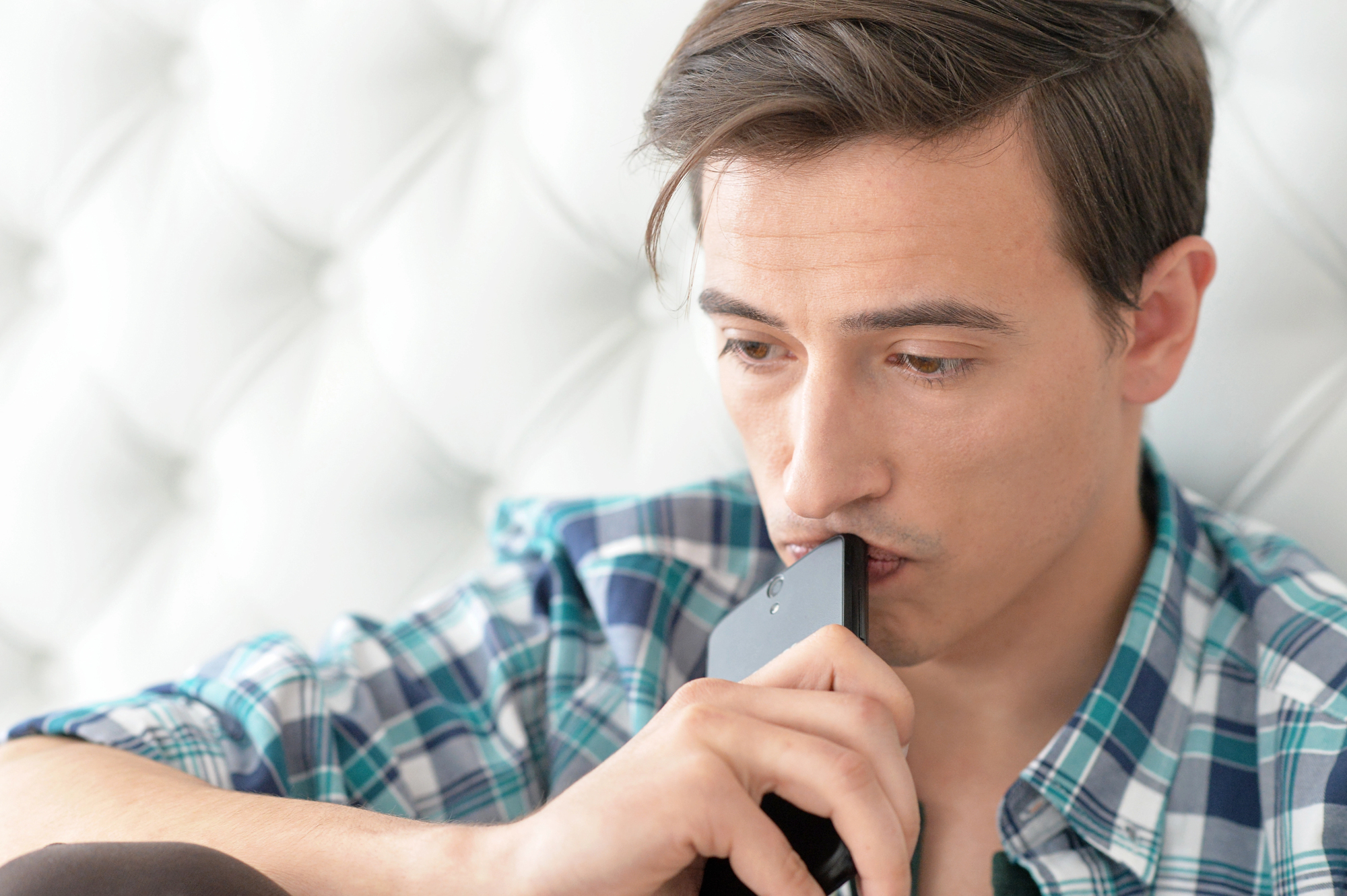 A young man with brown hair, wearing a blue and green checkered shirt, sits against a tufted white background. He holds a smartphone to his lips, looking thoughtful and concerned.