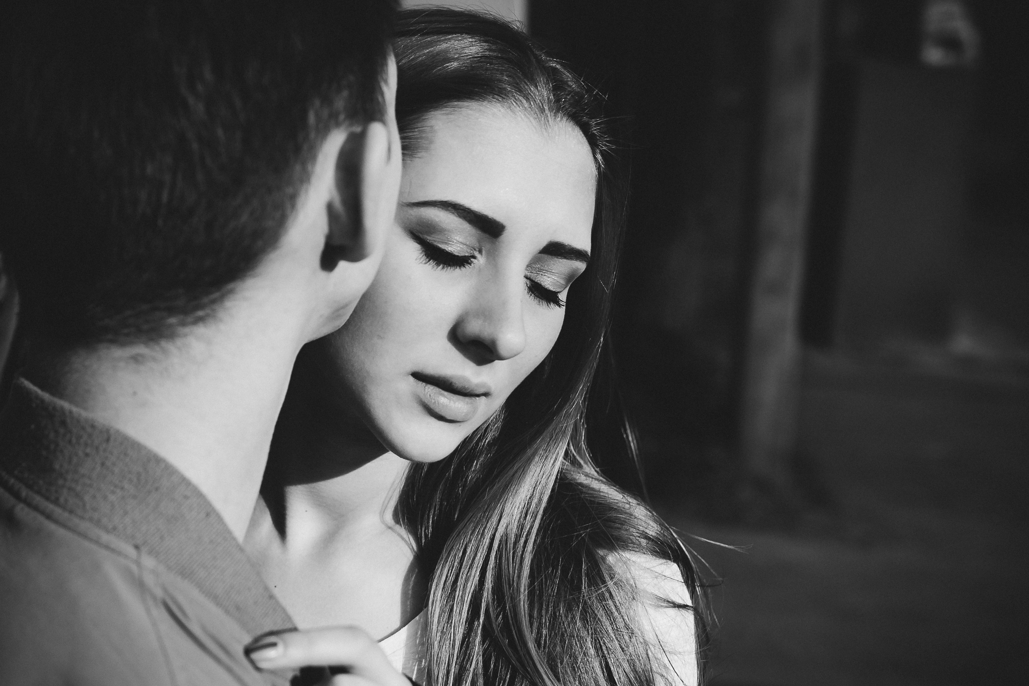 Black and white photo of a close-up of two people. One person, with long hair and closed eyes, appears calm and gently leans into the other person, who has their back to the camera. They are outside, with shadows cast on the ground in the background.