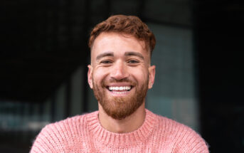 A man with short, curly brown hair and a beard is smiling broadly at the camera. He is wearing a pink knitted sweater and standing in front of a dark, blurred background.