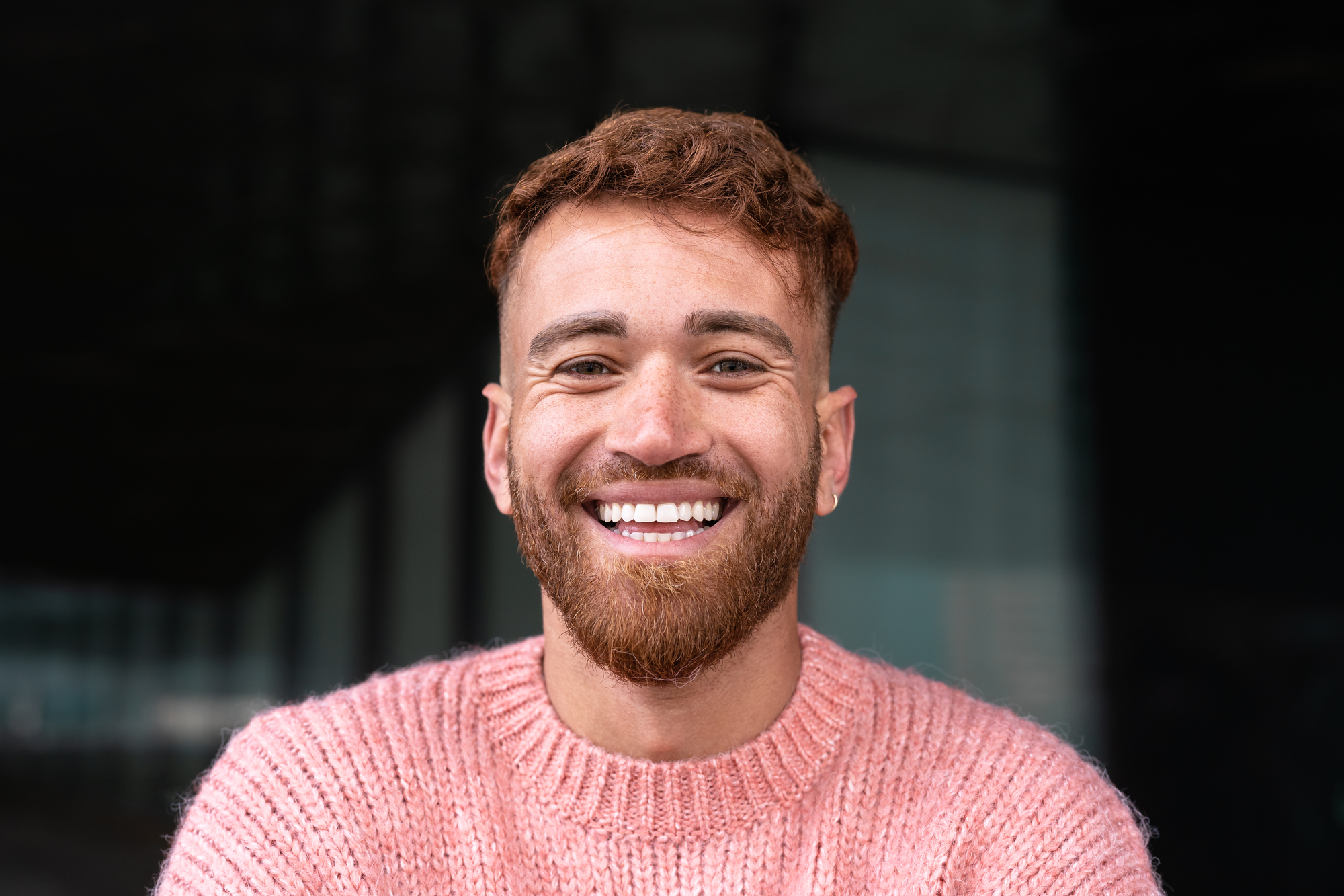 A man with short, curly brown hair and a beard is smiling broadly at the camera. He is wearing a pink knitted sweater and standing in front of a dark, blurred background.