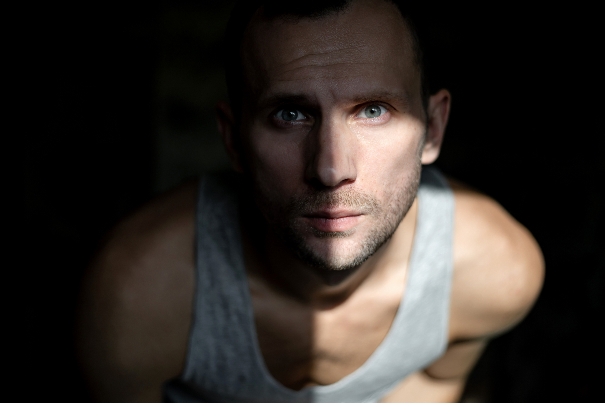 A man with a serious expression is looking directly at the camera. Light coming from the right side creates a dramatic shadow effect on his face. He is wearing a grey sleeveless shirt and has short hair. The background is dark and out of focus.