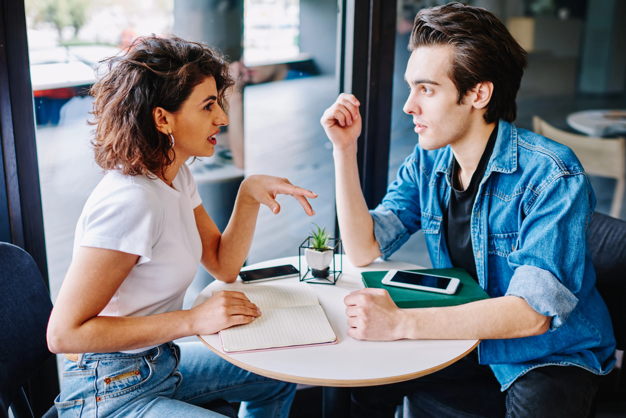 A woman with curly hair and a white shirt sits at a table with an open notebook, conversing with a man with styled hair and a denim jacket. Both have smartphones on the table, and a small potted plant is in the center. They appear to be in a modern café.