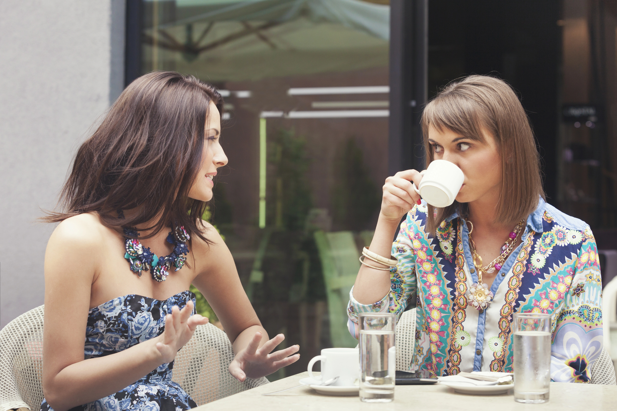 Two women sit at an outdoor cafe table, engaged in conversation. One woman, wearing a floral dress and statement necklace, is talking animatedly with gestures. The other woman, in a colorful patterned shirt, is holding a white coffee cup near her mouth.