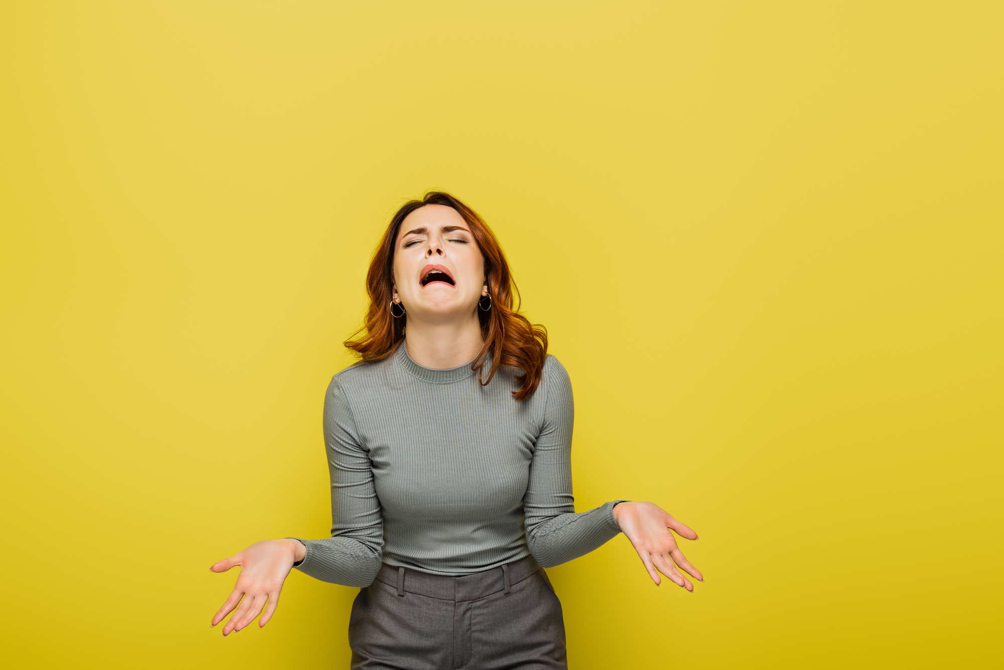 A woman stands in front of a yellow background with her arms outstretched and an upset facial expression. She is wearing a long-sleeve gray top and gray pants, conveying a sense of frustration or distress.