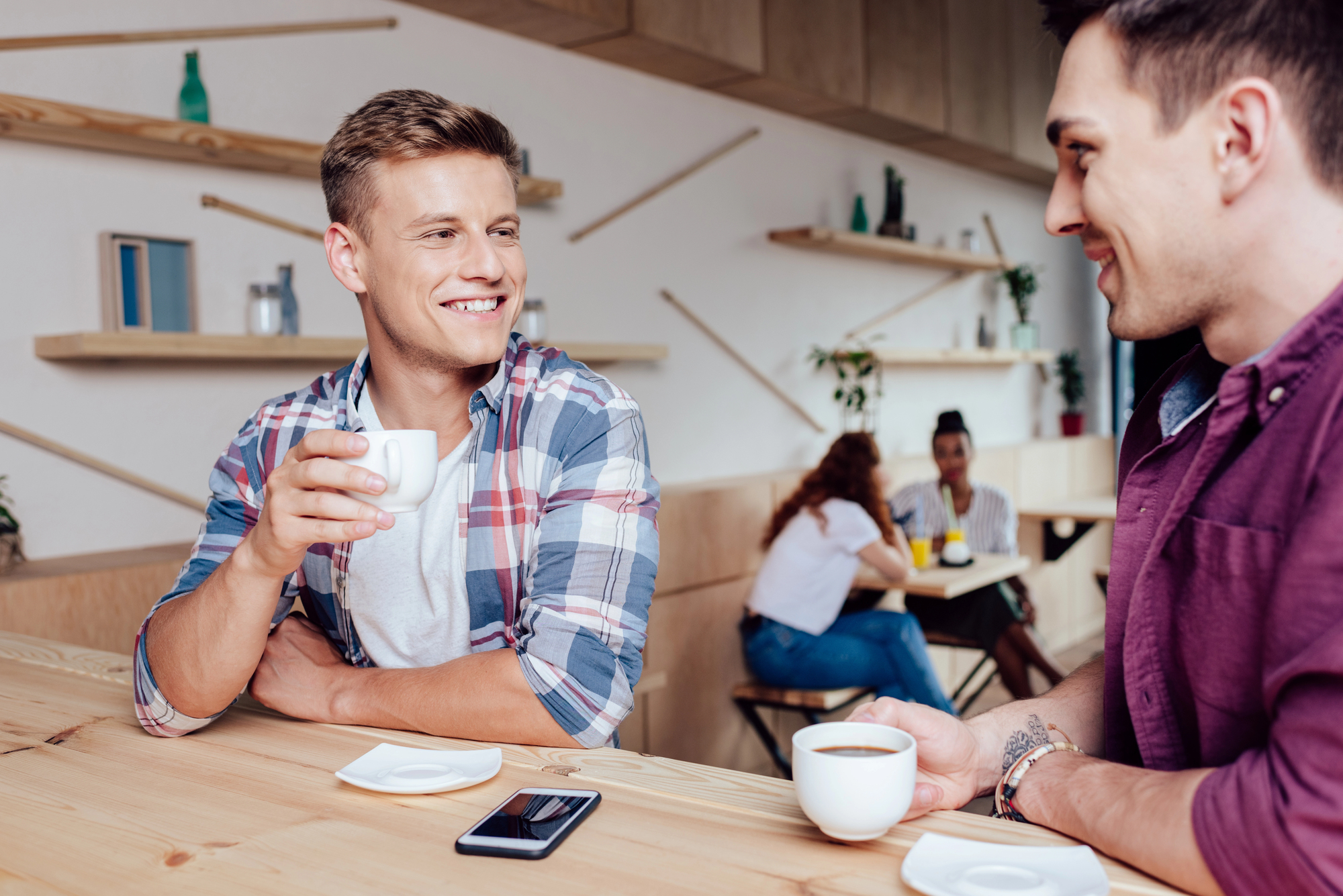 Two men enjoying coffee together at a modern café. One man, wearing a plaid shirt, holds his cup mid-conversation and smiles at the other man, who wears a maroon shirt and also has a coffee cup in hand. A smartphone lies on the table between them.