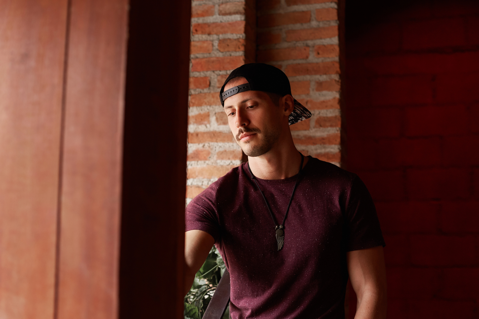 A person wearing a backwards baseball cap, a maroon t-shirt, and a necklace with a pendant stands near a wooden beam and a brick wall, looking down pensively. The person has short hair and a trimmed mustache.