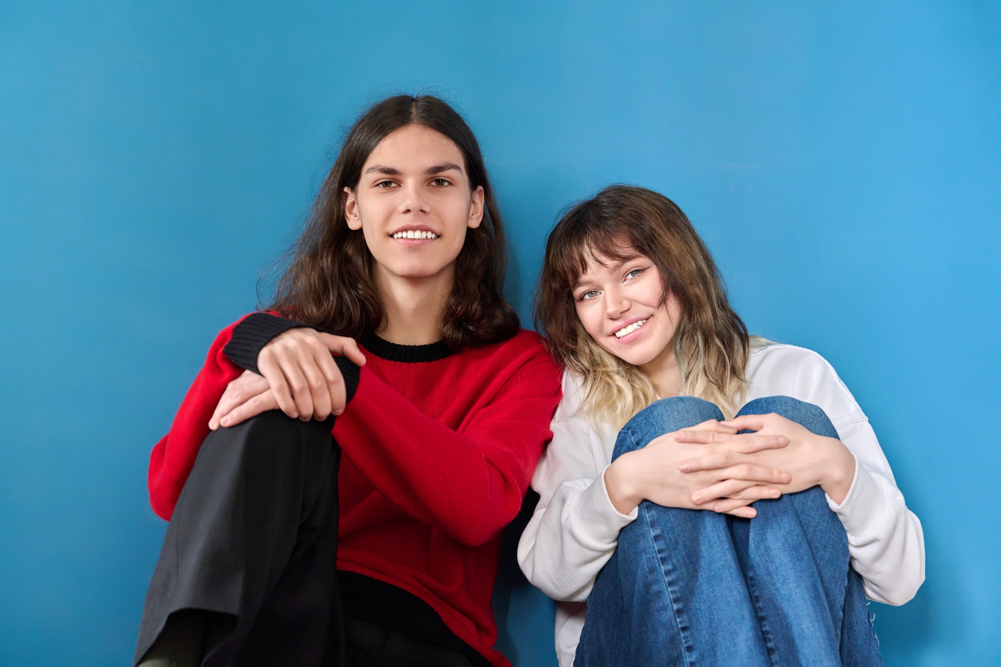 Two young people sit against a blue background, smiling at the camera. The person on the left has long dark hair, wears a red sweater, and black pants. The person on the right has shoulder-length light brown hair with bangs, wears a white shirt and blue jeans.