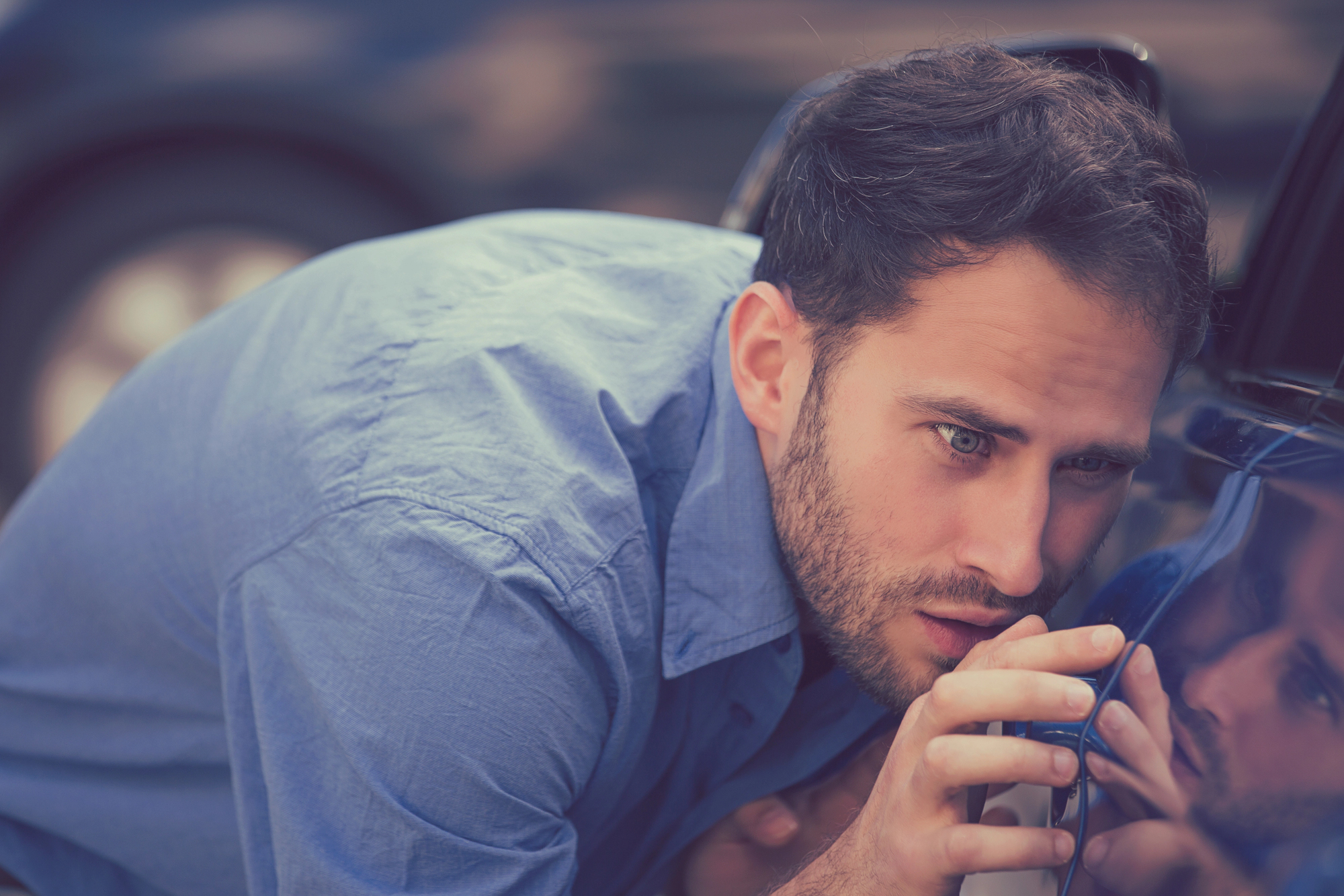 A man in a blue shirt is closely inspecting the side mirror of a car, with a focused expression. The blue car and the man's reflection are clearly visible, suggesting he is carefully examining or repairing the vehicle.