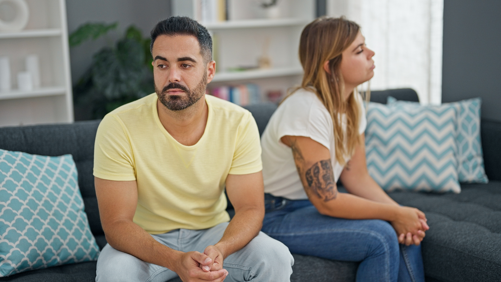 A man and a woman sit on a couch looking away from each other. The man, with dark hair and a beard, wears a yellow shirt. The woman, with light brown hair, wears a white shirt and jeans, showing a tattoo on her left arm. The room has a grey couch and patterned cushions.