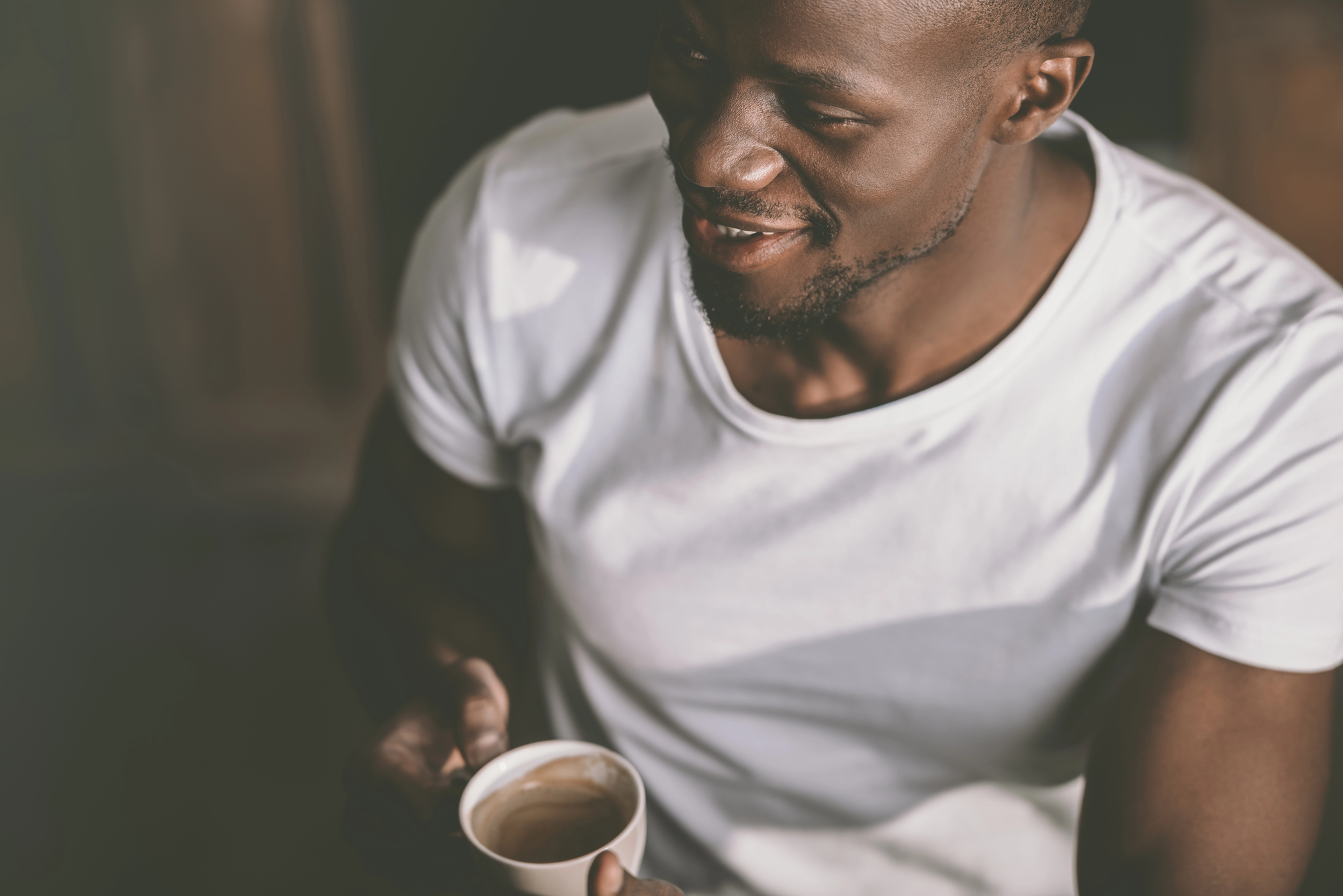 A man wearing a white t-shirt is holding a small cup of coffee and smiling. The background is dimly lit.