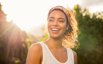 A woman with curly hair wearing a headband and a sleeveless top smiles brightly in the sunlight. She is outdoors with greenery in the background, and the sun creates a warm glow around her.