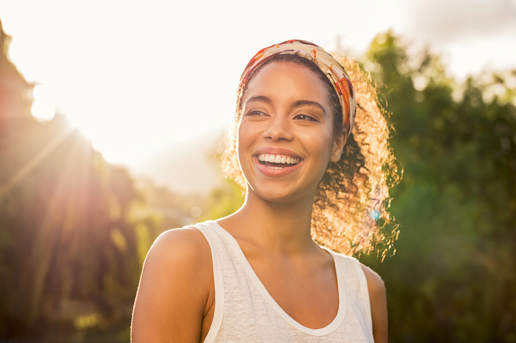 A woman with curly hair wearing a headband and a sleeveless top smiles brightly in the sunlight. She is outdoors with greenery in the background, and the sun creates a warm glow around her.