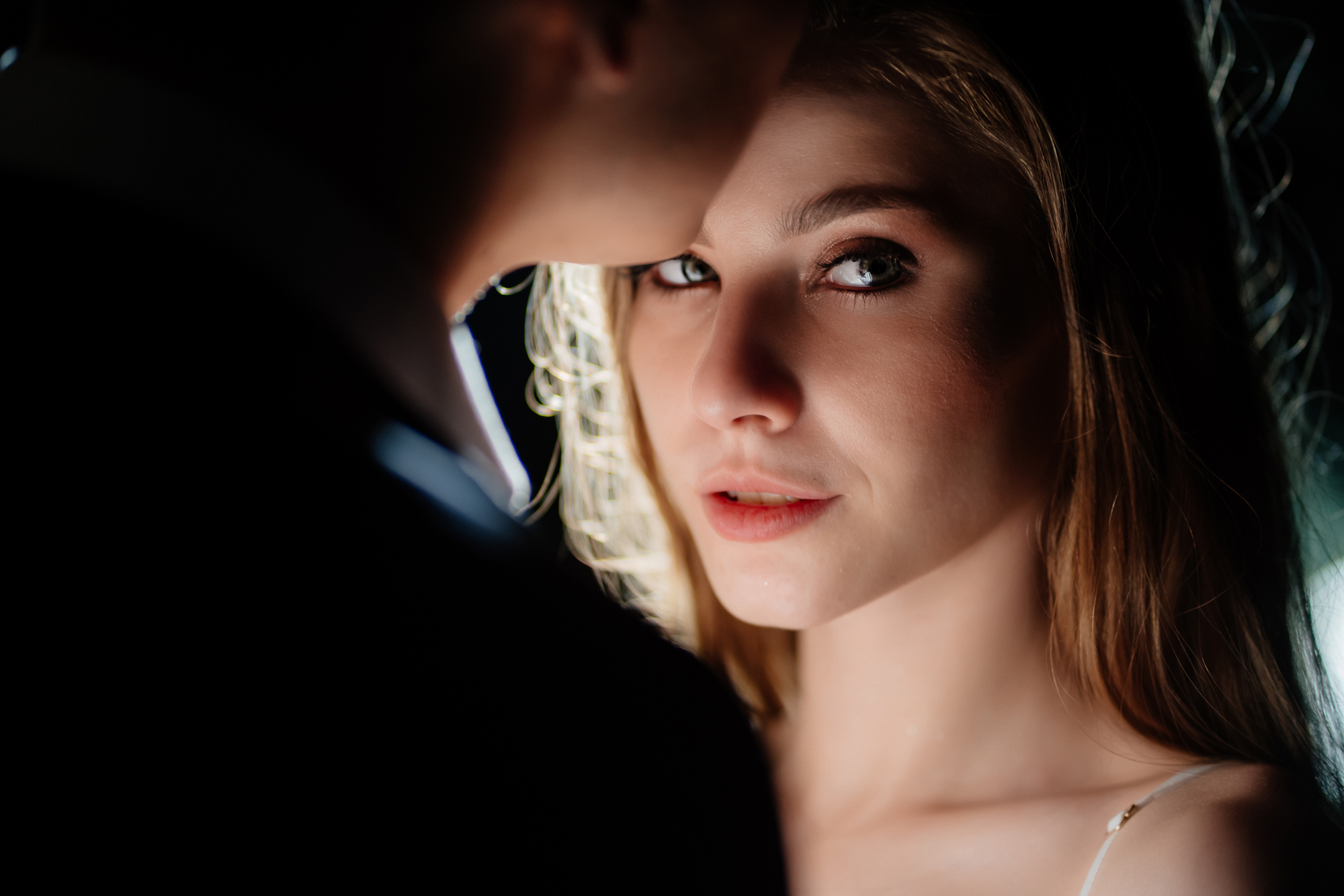 A close-up shot of a woman with long blonde hair and light skin, looking directly at the camera with a neutral expression. She is partially obscured by a man next to her, whose face is not fully visible. The background is dark and the lighting is dramatic.