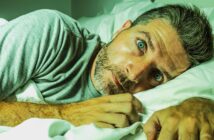 A man with short, graying hair and a beard lies under white sheets in bed, wearing a gray t-shirt. He looks directly at the camera with wide, blue eyes, appearing contemplative or startled. The lighting casts a greenish tint on the image.