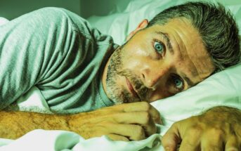 A man with short, graying hair and a beard lies under white sheets in bed, wearing a gray t-shirt. He looks directly at the camera with wide, blue eyes, appearing contemplative or startled. The lighting casts a greenish tint on the image.