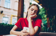 A woman in a red shirt sits at an outdoor cafe, resting her head on her hand and looking down at her smartphone with a pensive expression. There are blurred buildings and greenery in the background.