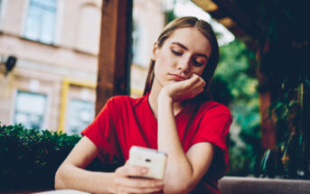 A woman in a red shirt sits at an outdoor cafe, resting her head on her hand and looking down at her smartphone with a pensive expression. There are blurred buildings and greenery in the background.