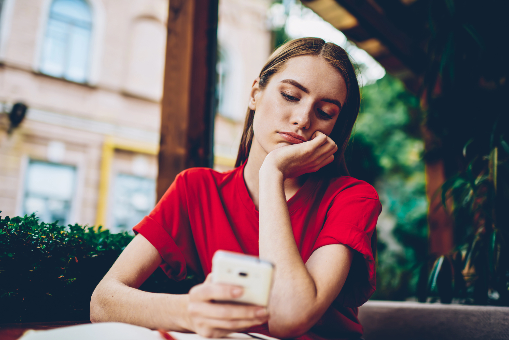 A woman in a red shirt sits at an outdoor cafe, resting her head on her hand and looking down at her smartphone with a pensive expression. There are blurred buildings and greenery in the background.