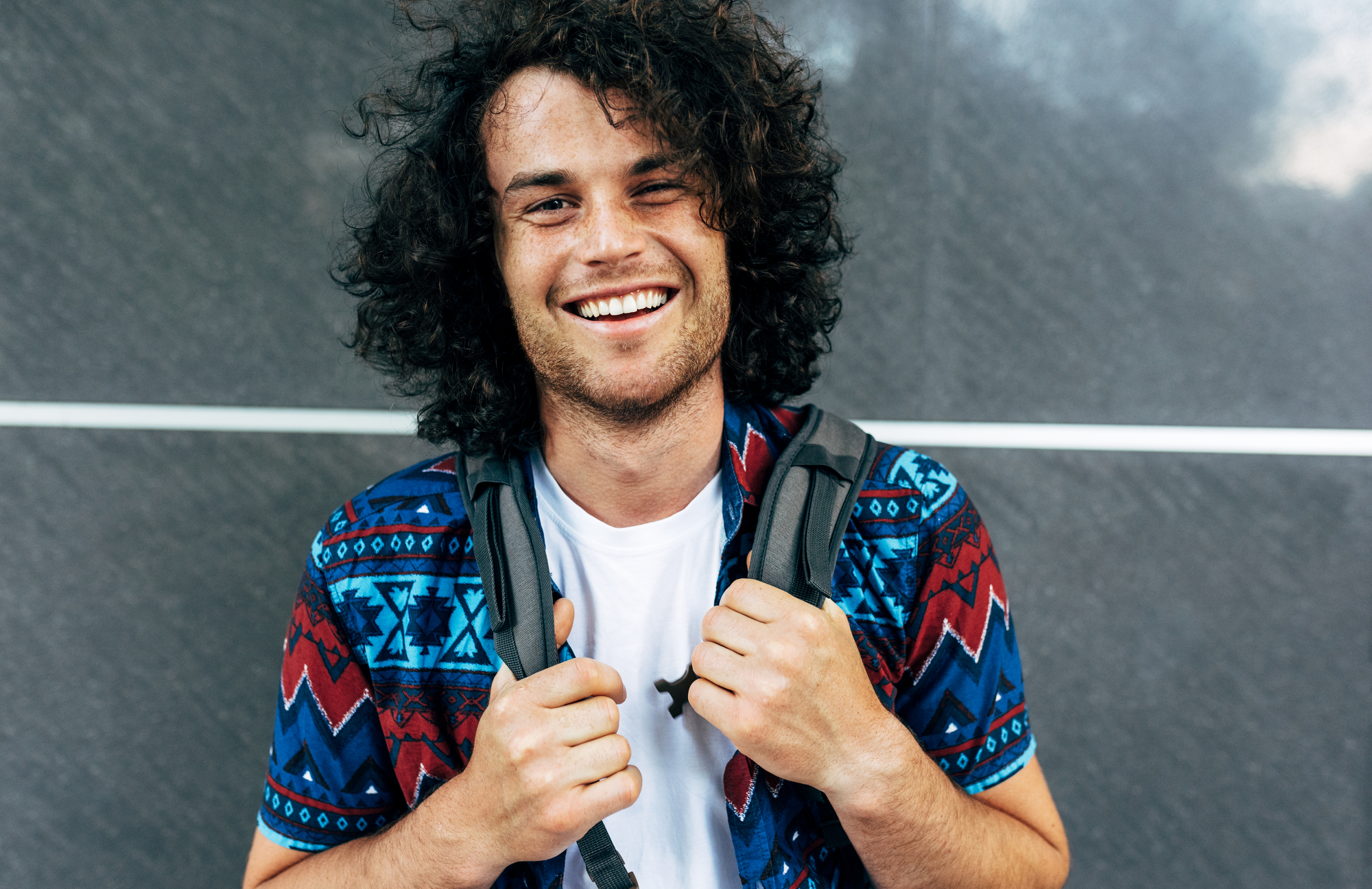 A man with curly hair smiles while holding the straps of his backpack. He wears a colorful patterned shirt over a white t-shirt and stands against a dark gray wall background.