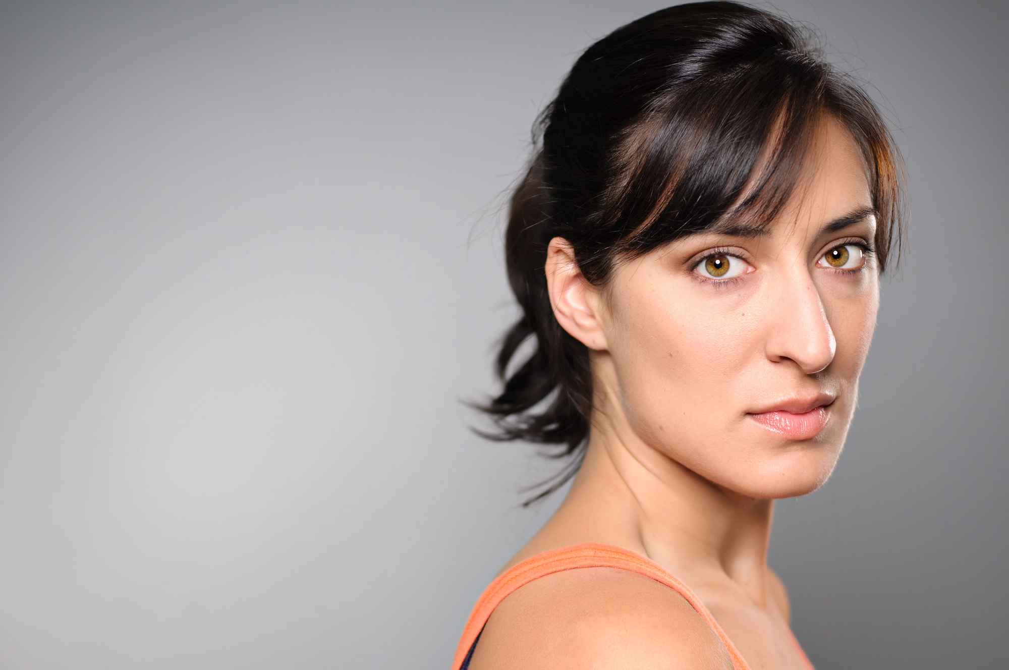 A woman with dark hair pulled back looks towards the camera with a neutral expression. She is wearing a sleeveless orange top against a plain grey background.